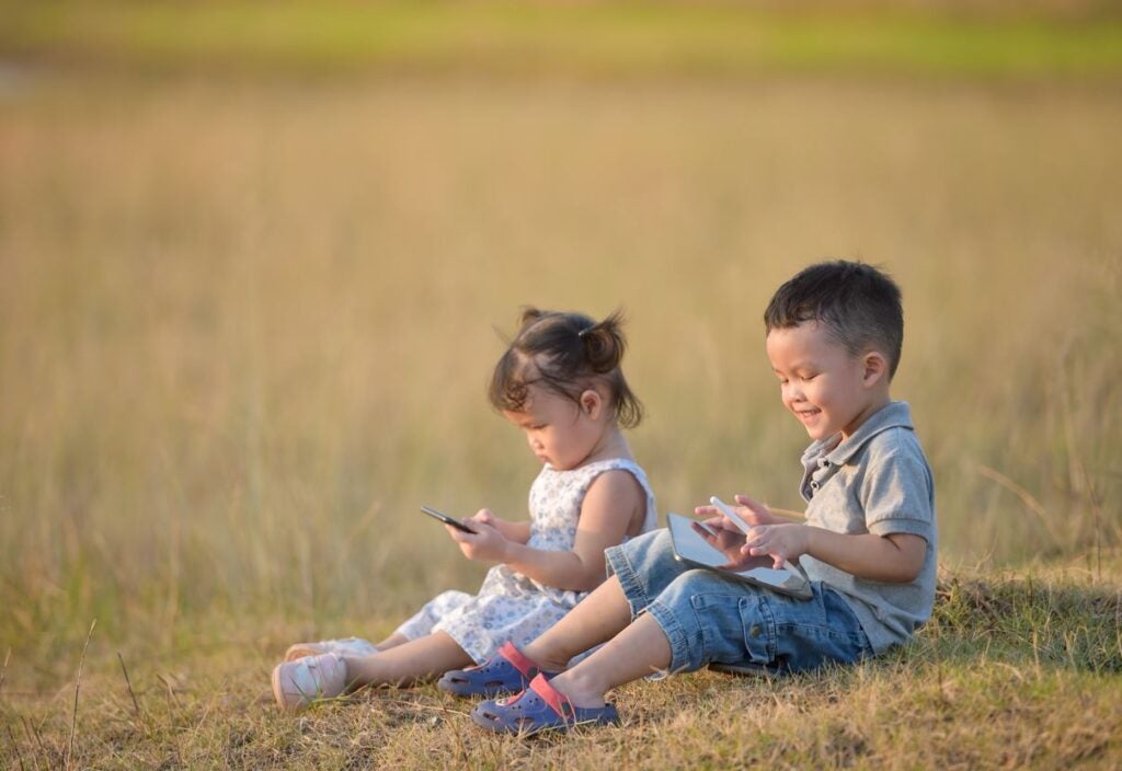 Una niña usando celular y un niño usando tableta en un campo abierto