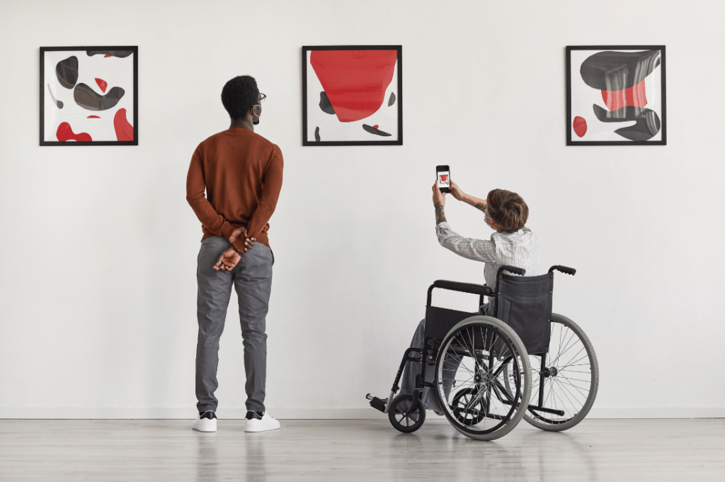 Joven en silla de ruedas fotografía una pintura moderna en un museo, acompañado de un amigo.