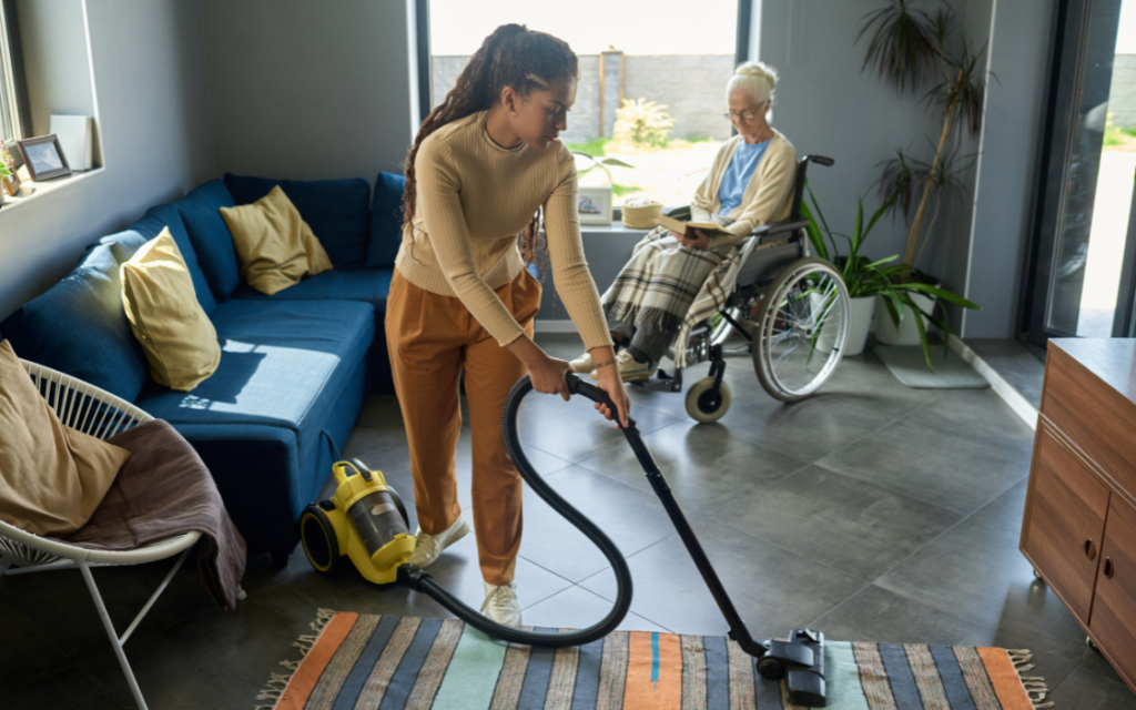 Mujer joven limpia alfombra con la aspiradora mientras cuida de adulta mayor en silla de ruedas