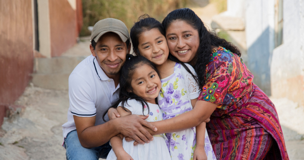 Familia latinoamericana conformada por papá, mamá y dos hijas sonriendo abrazados.