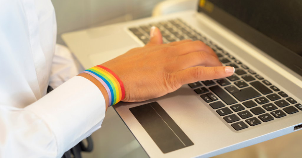 Manos de una persona escribiendo en una laptop, mientras usa un brazalete con la bandera LGBTQ+