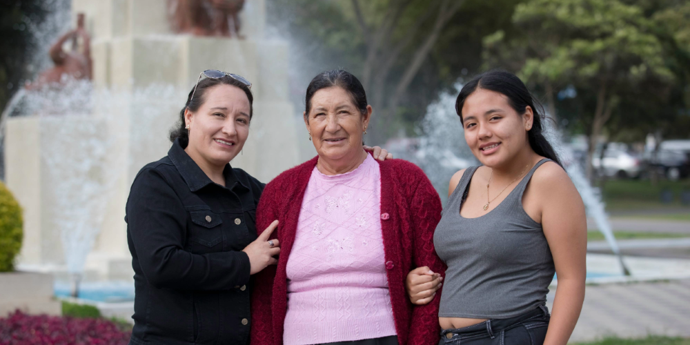 Abuela, madre e hija latinoamericanas sonriendo a la cámara en una plaza.