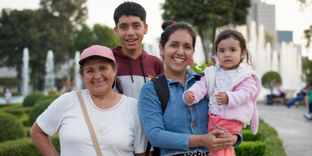 Una familia compuesta de abuela, madre, hijo mayor e hija menor sonriendo a la cámara en una ciudad latinoamericana.