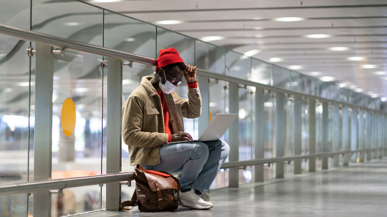 La imagen muestra un joven en un aeropuerto utilizando mascarilla teletrabajando